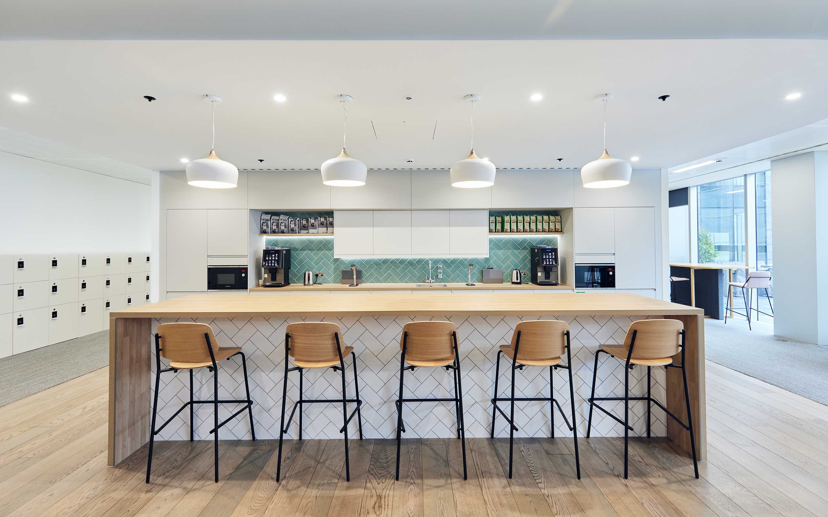 The image is of an office teapoint. High wooden breakfast stools tuck under an island unit. The floor is wood, and the kitchen backsplash tiles are duck egg blue
