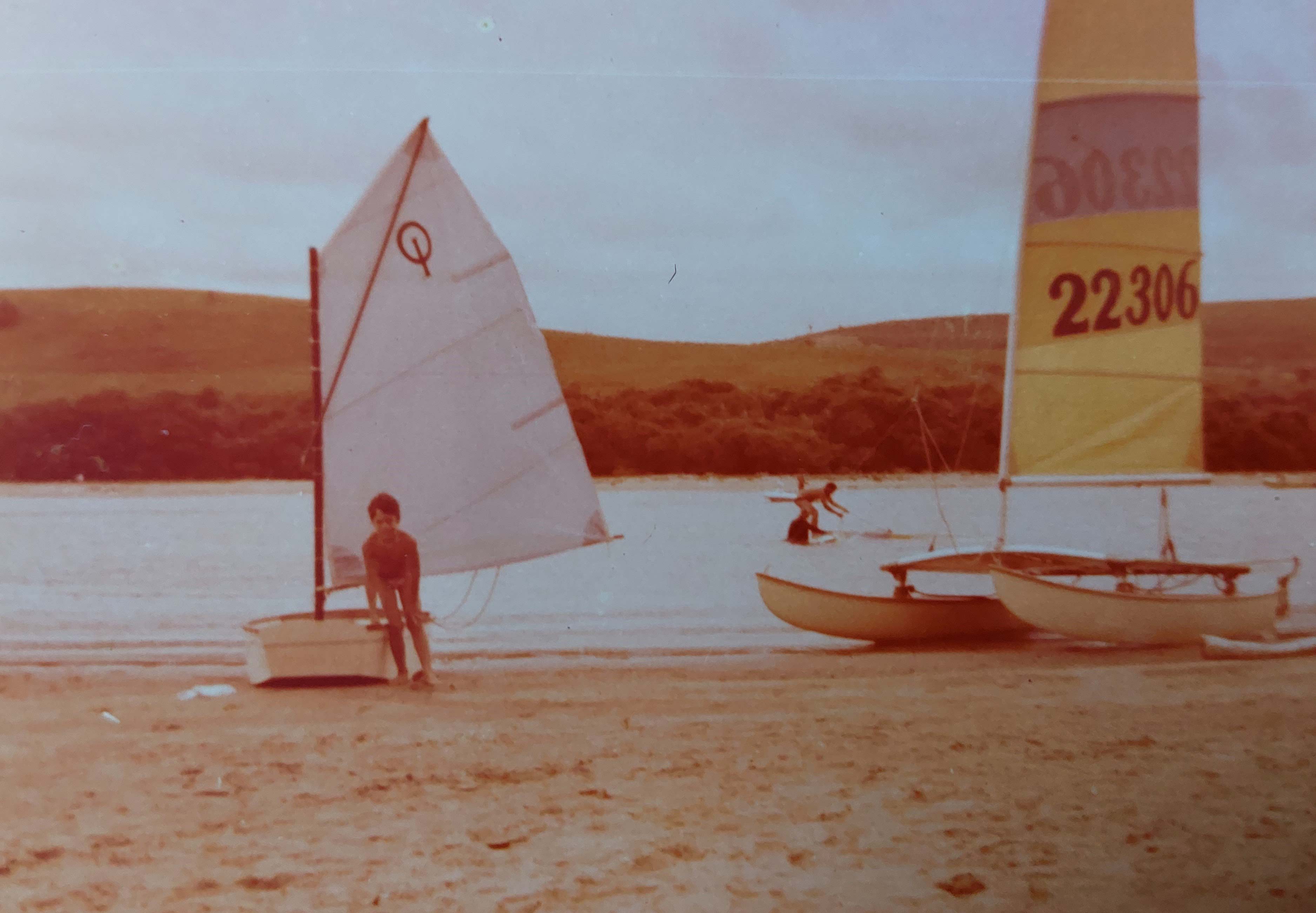 A young boy standing next to some sail boats on a beach. The boats have white and yellow sails.
