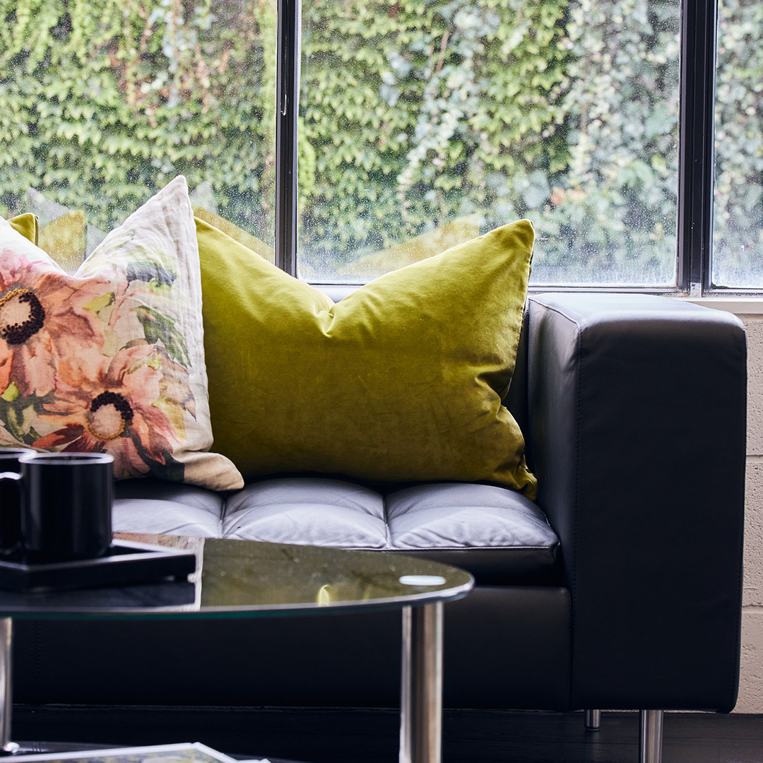 The image is a detailed shot of green and floral sofa cushions, on a dark leather sofa, with a window behind