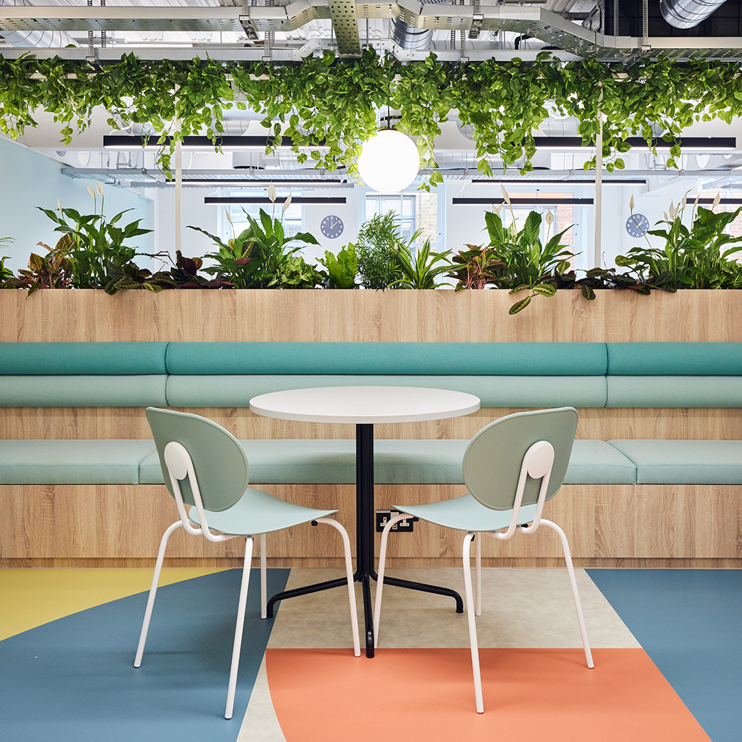 The image shows a cafeteria table, chairs and banquette seating, integrated with greenery and office plants and a colourful floor