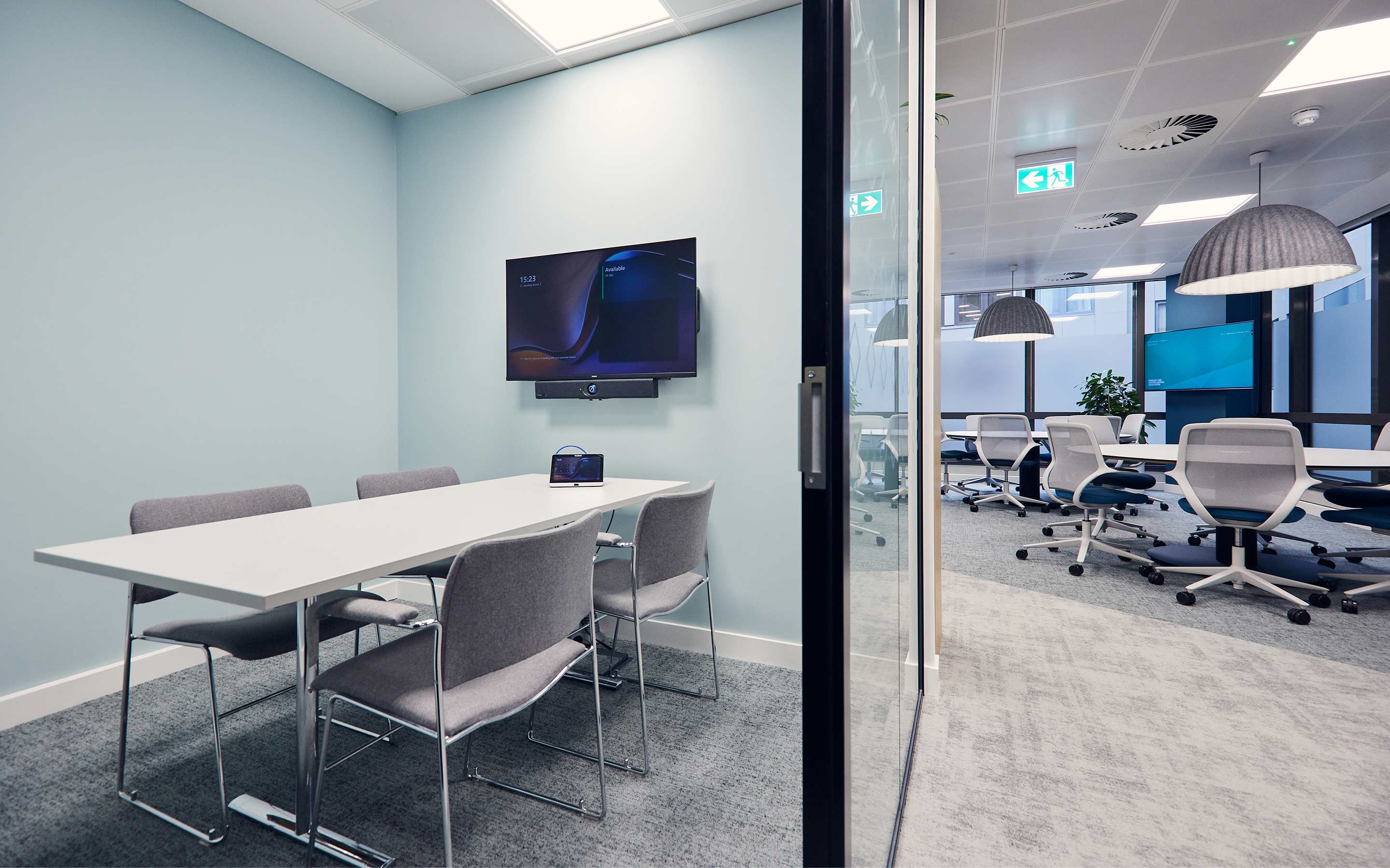 The image shows both a meeting room with glass walls, and a breakout collaboration area beyond with large pendant lighting and round tables