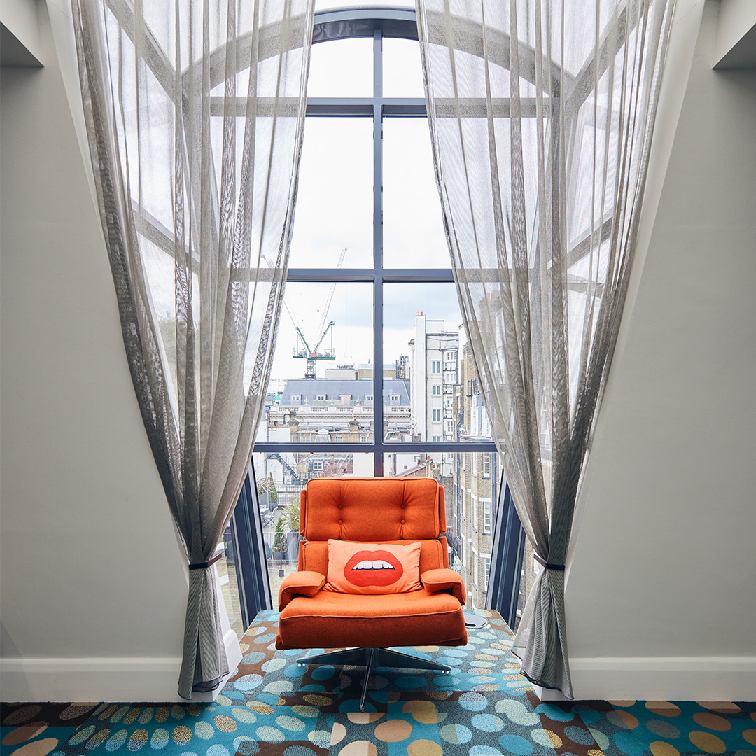 The image shows an orange chair framed in a window with sheer curtains and views of the city of london