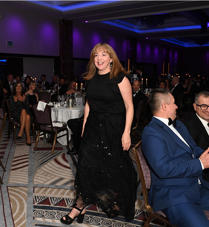 A person in a black dress at a formal event with seated guests and dinner tables.