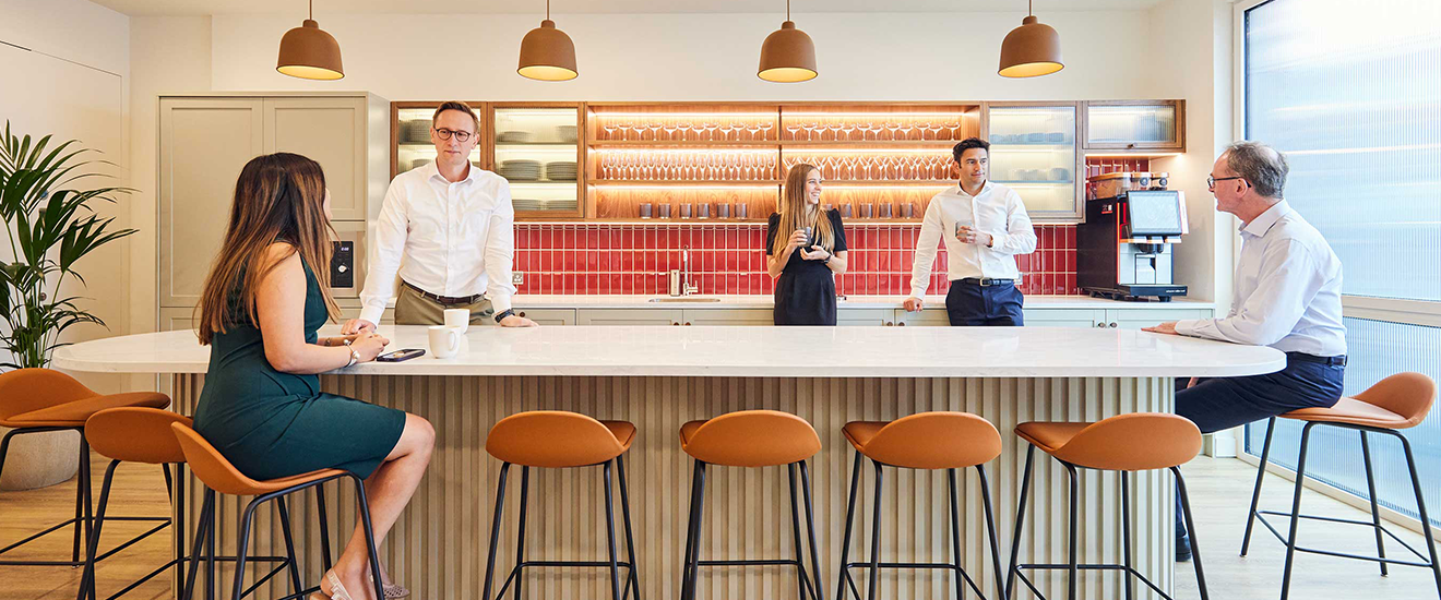 Modern office kitchen area with employees around a white bar table, pendant lights, and red backsplash.