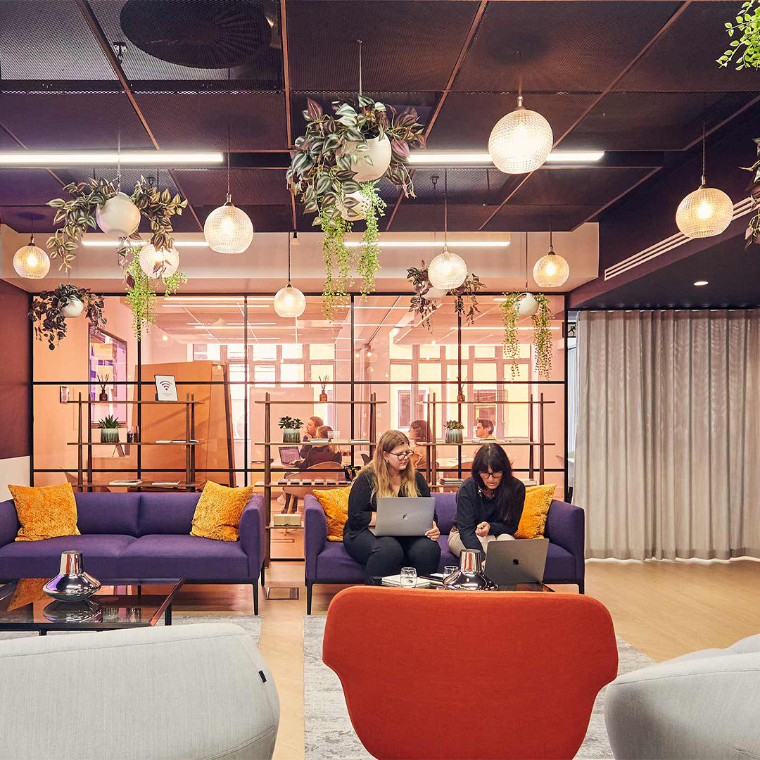 2 women sat on a purple sofa with yellow cushions looking at laptops discussing work.