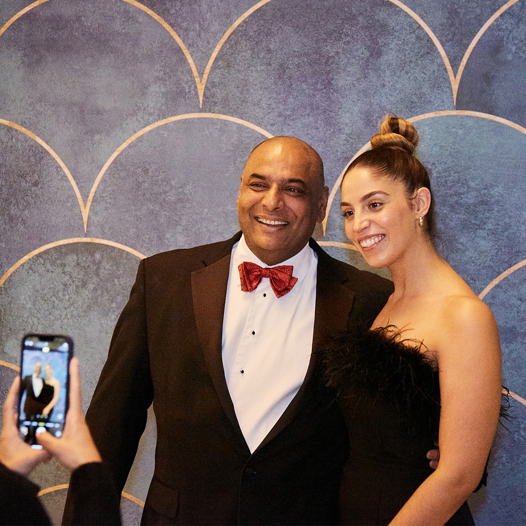 A man and woman smiling together for a photo at a social event, dressed elegantly and enjoying the moment.