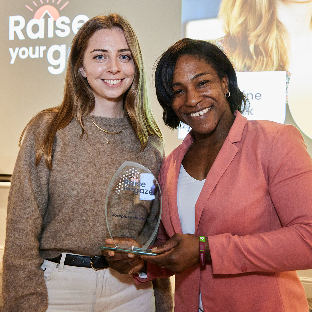 In front of a screen, two women hold an award, showcasing their accomplishment and camaraderie in a celebratory moment