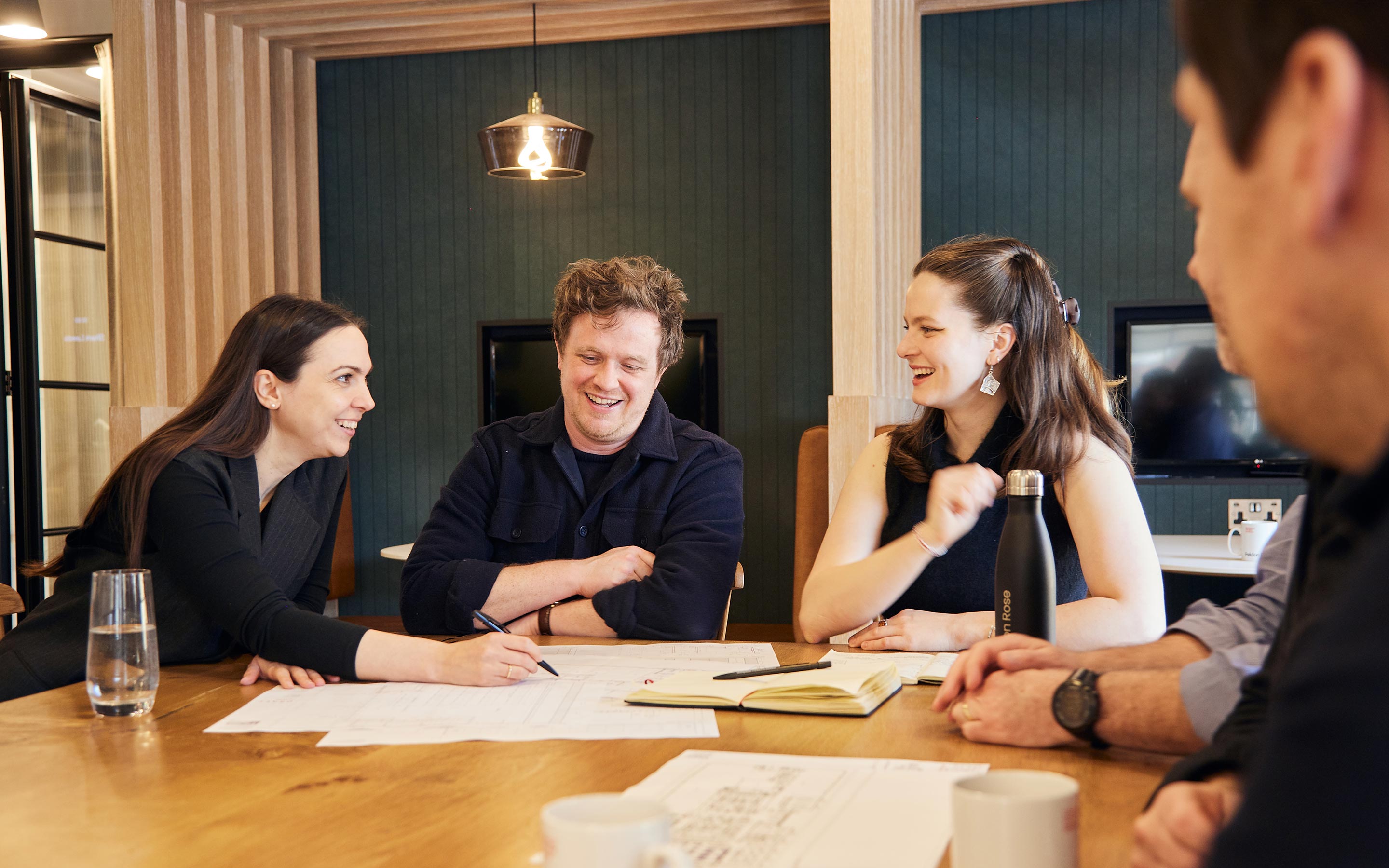 Colleagues sit jovially around a kitchen table in a modern office, collaborating and enjoying each other's company