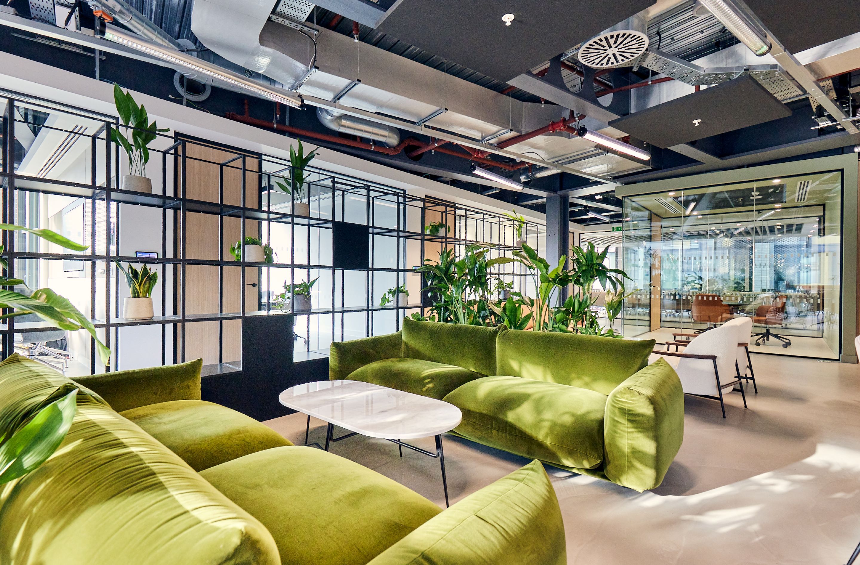 Modern office lounge area with green sofas, plants, and glass partition.