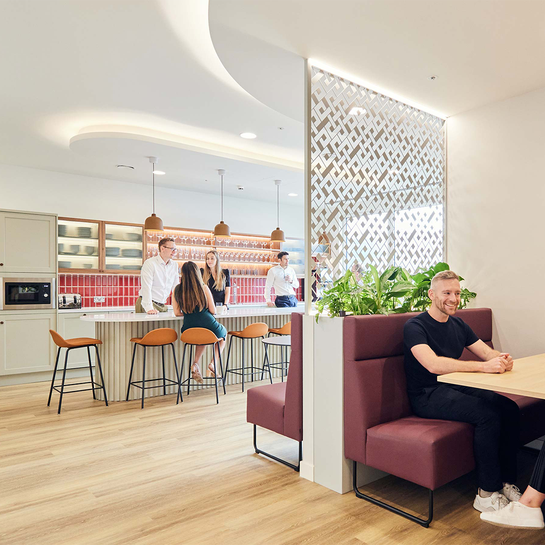 A man sad on a purple sofa in a workspace booth smiling talking to someone, Infront of a kitchen bar with people standing around chatting.