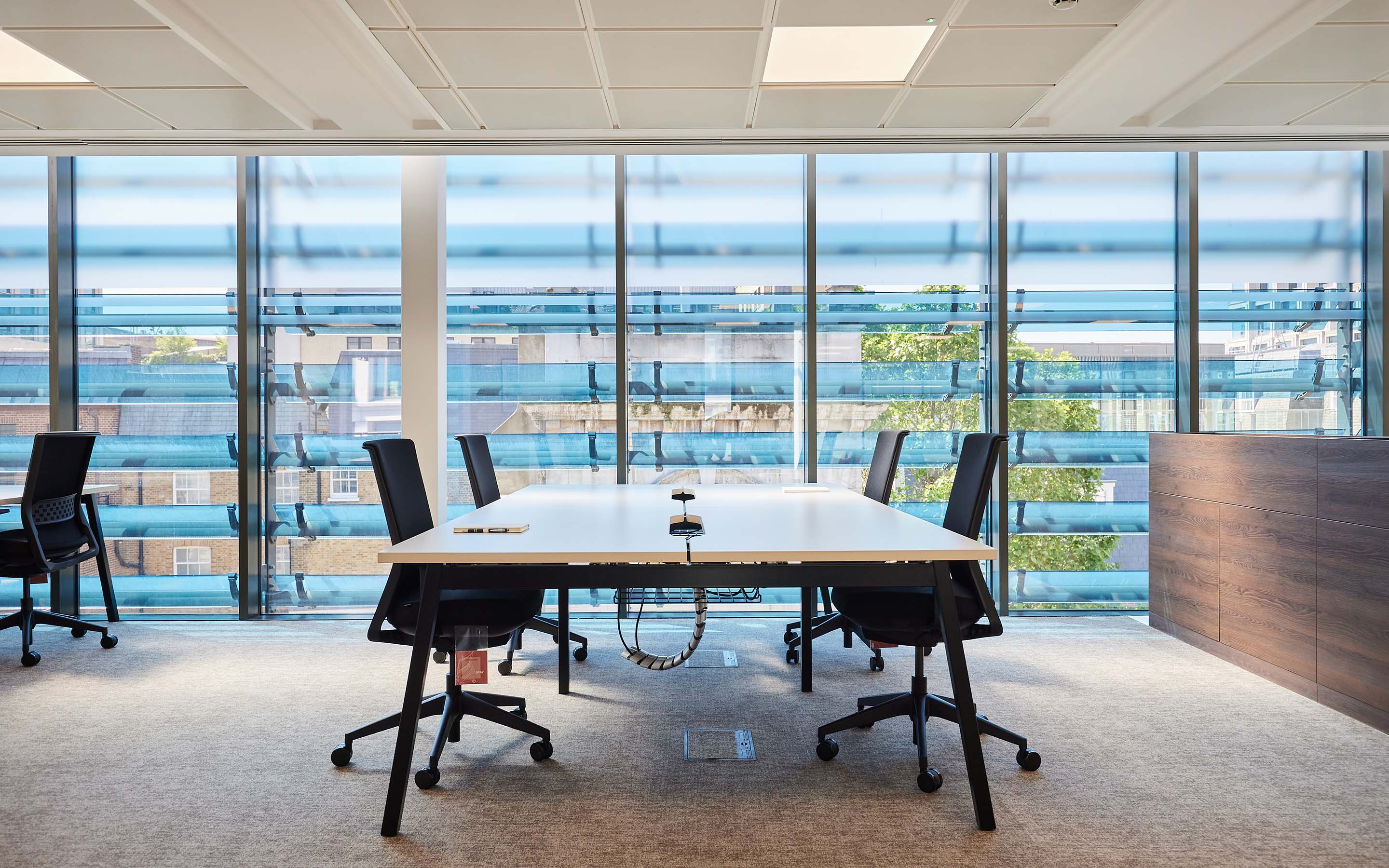 Modern office meeting room with long white table, black chairs, and large windows offering city views.