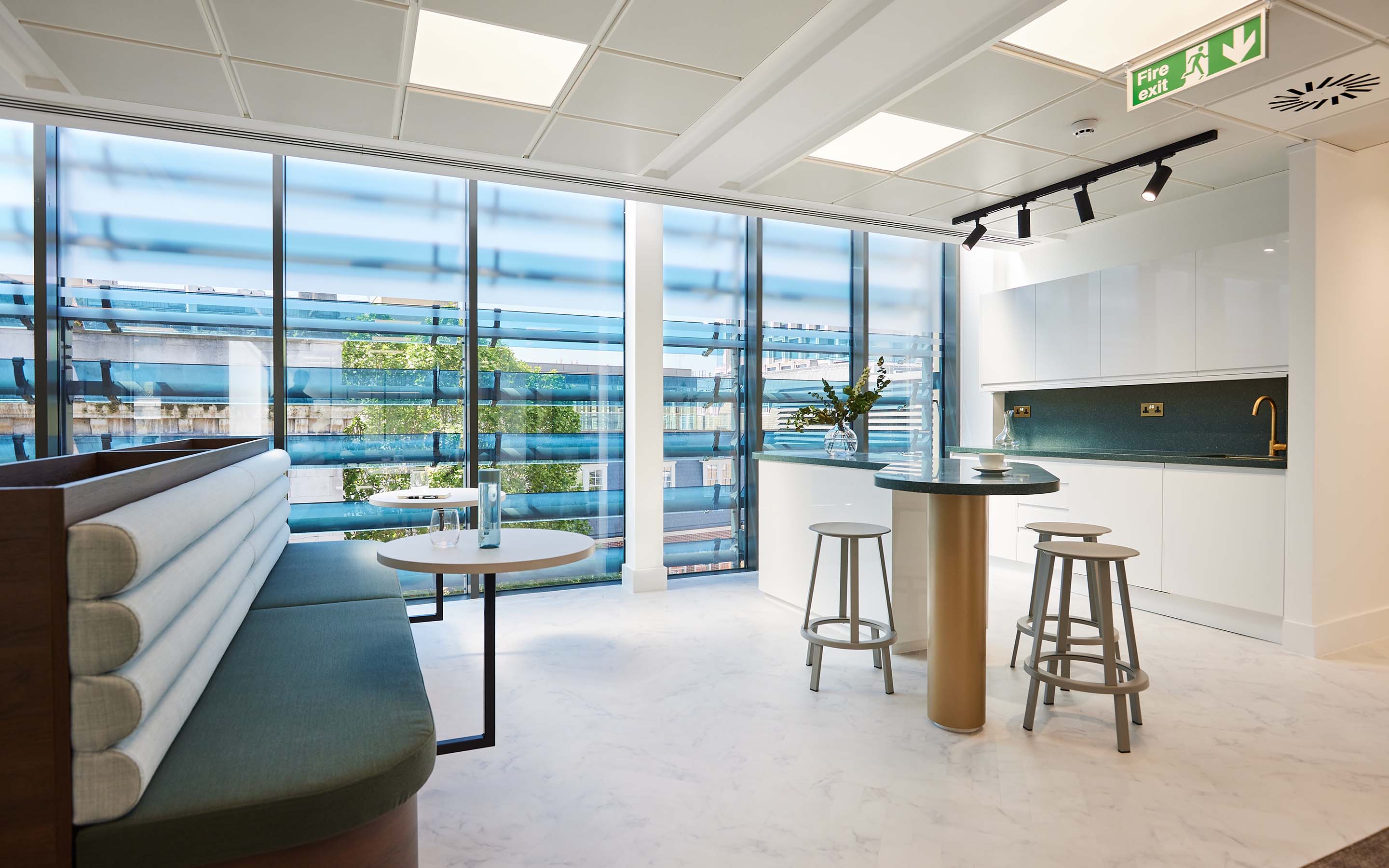 Modern office break area with seating, table, stools, and large windows overlooking urban scenery.