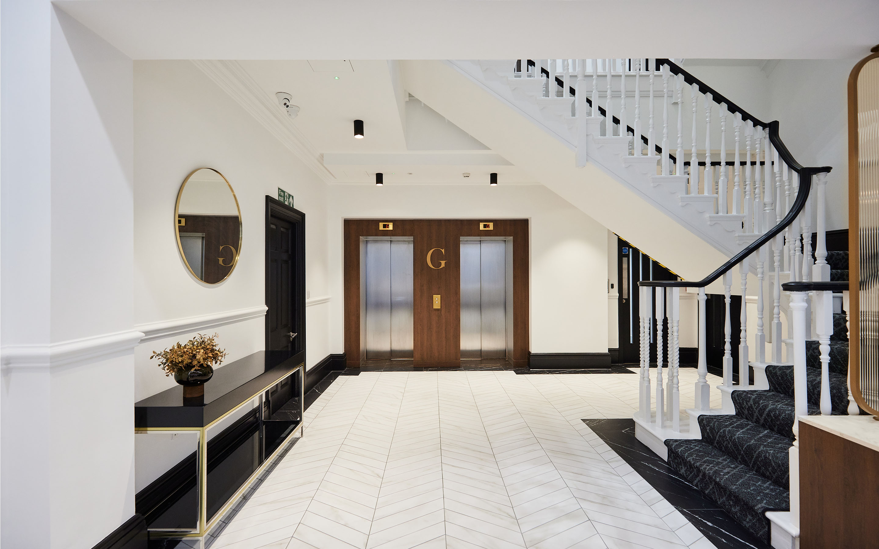 Elegant interior with white staircase, black-trimmed elevator doors marked with a 'G', and herringbone floor.