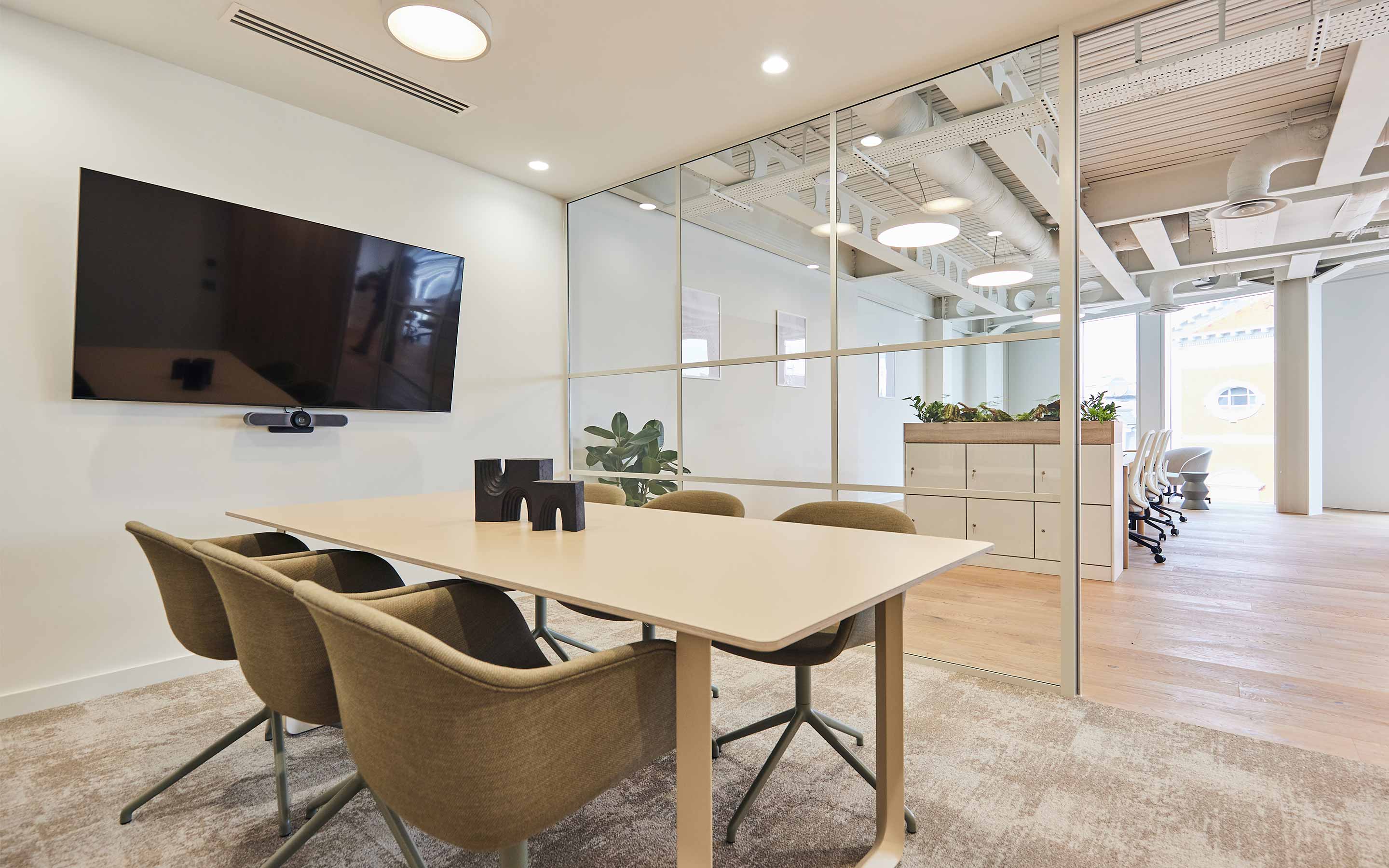 A meeting room in a modern office, with soft seating, a wall mounted TV screen, and glass walls