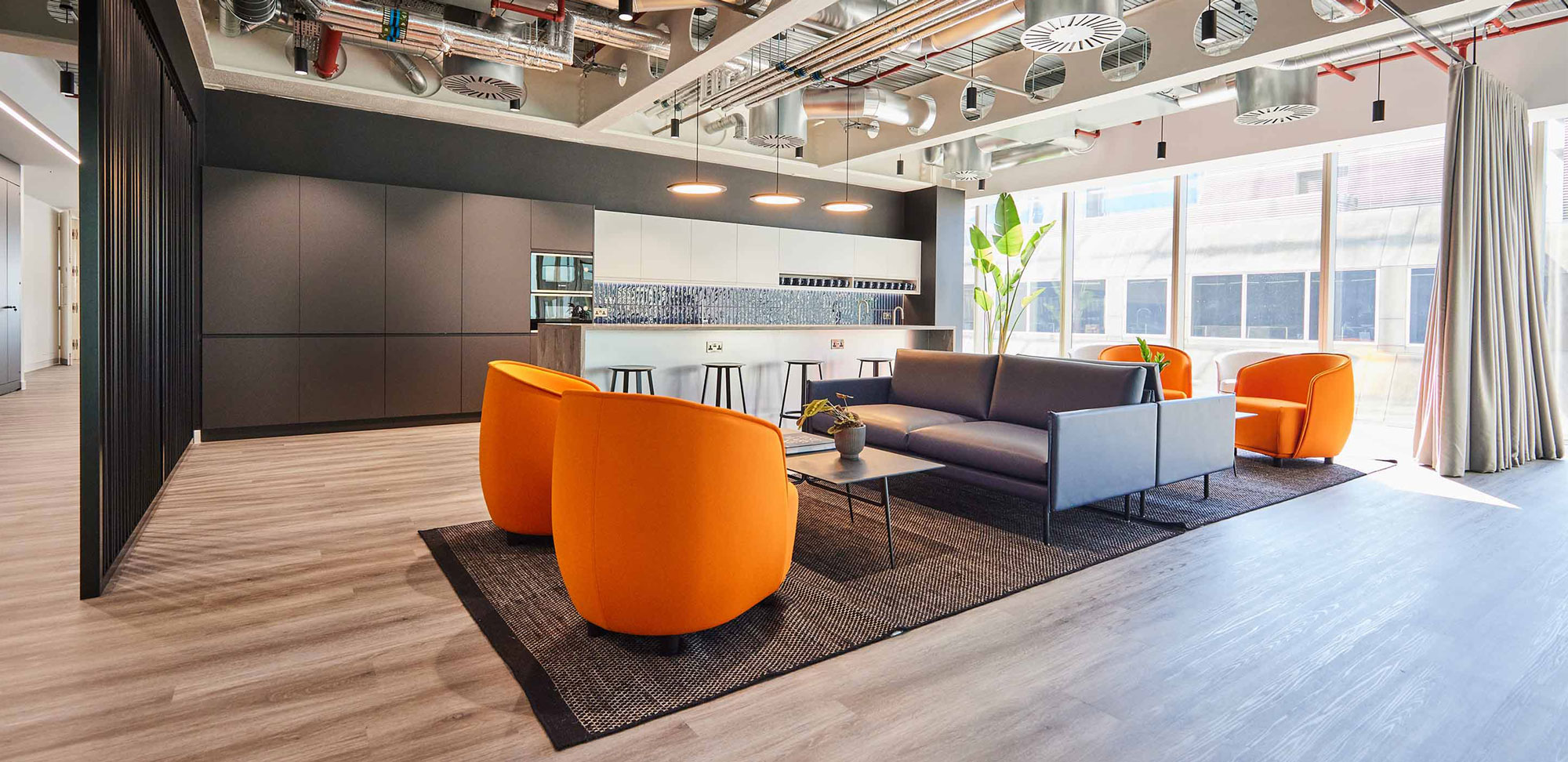 Modern office lounge with orange chairs, a gray sofa, and kitchen area.