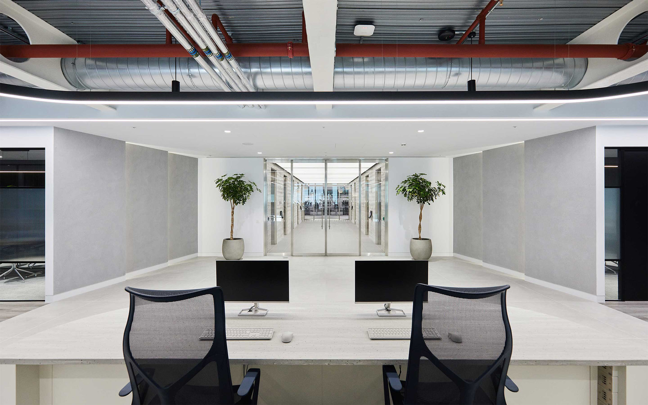 Modern office reception area with two monitors on a desk, plant decor, and visible ceiling pipes.