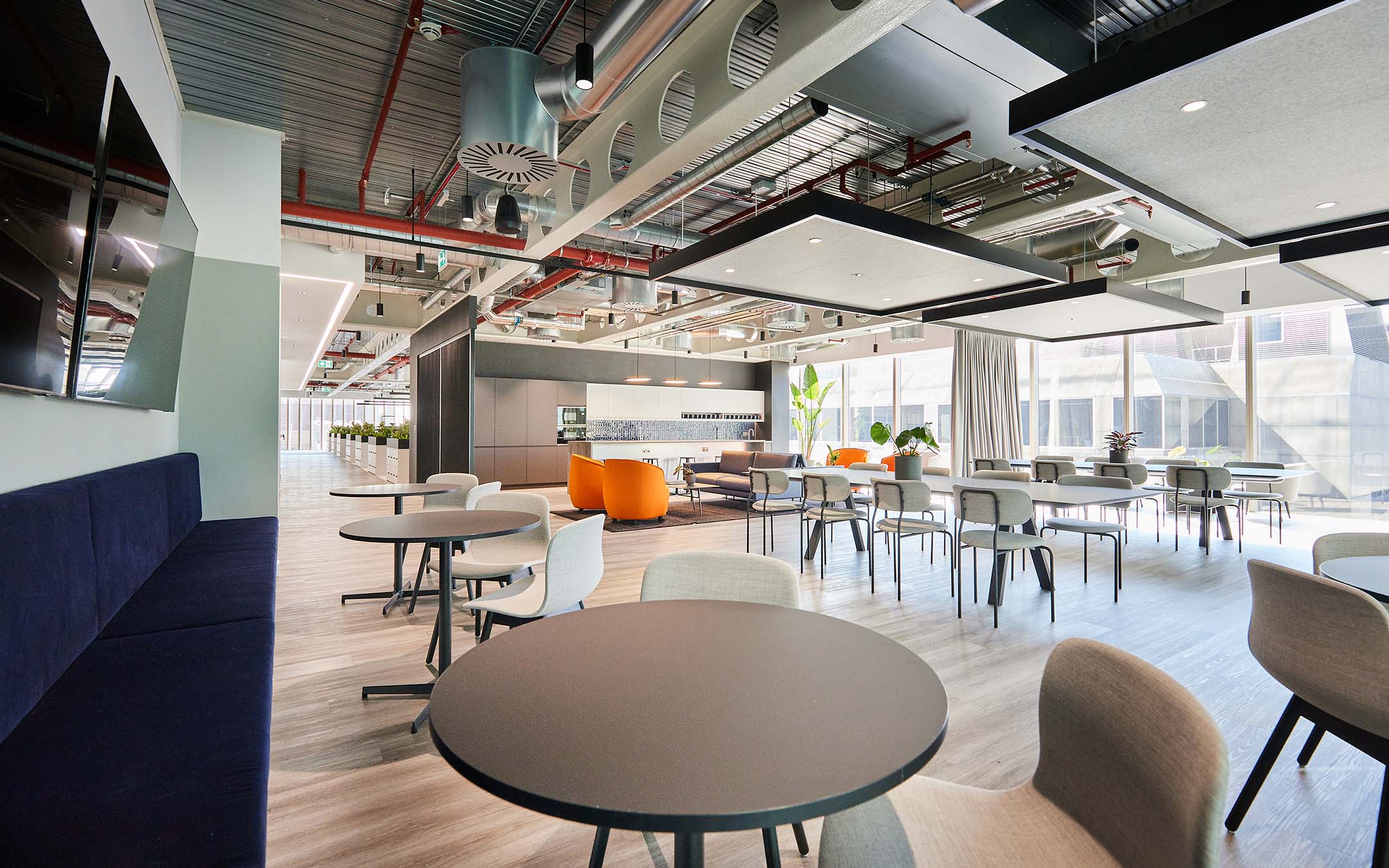 Modern office break room with tables, chairs, and lounge area under exposed ceiling.