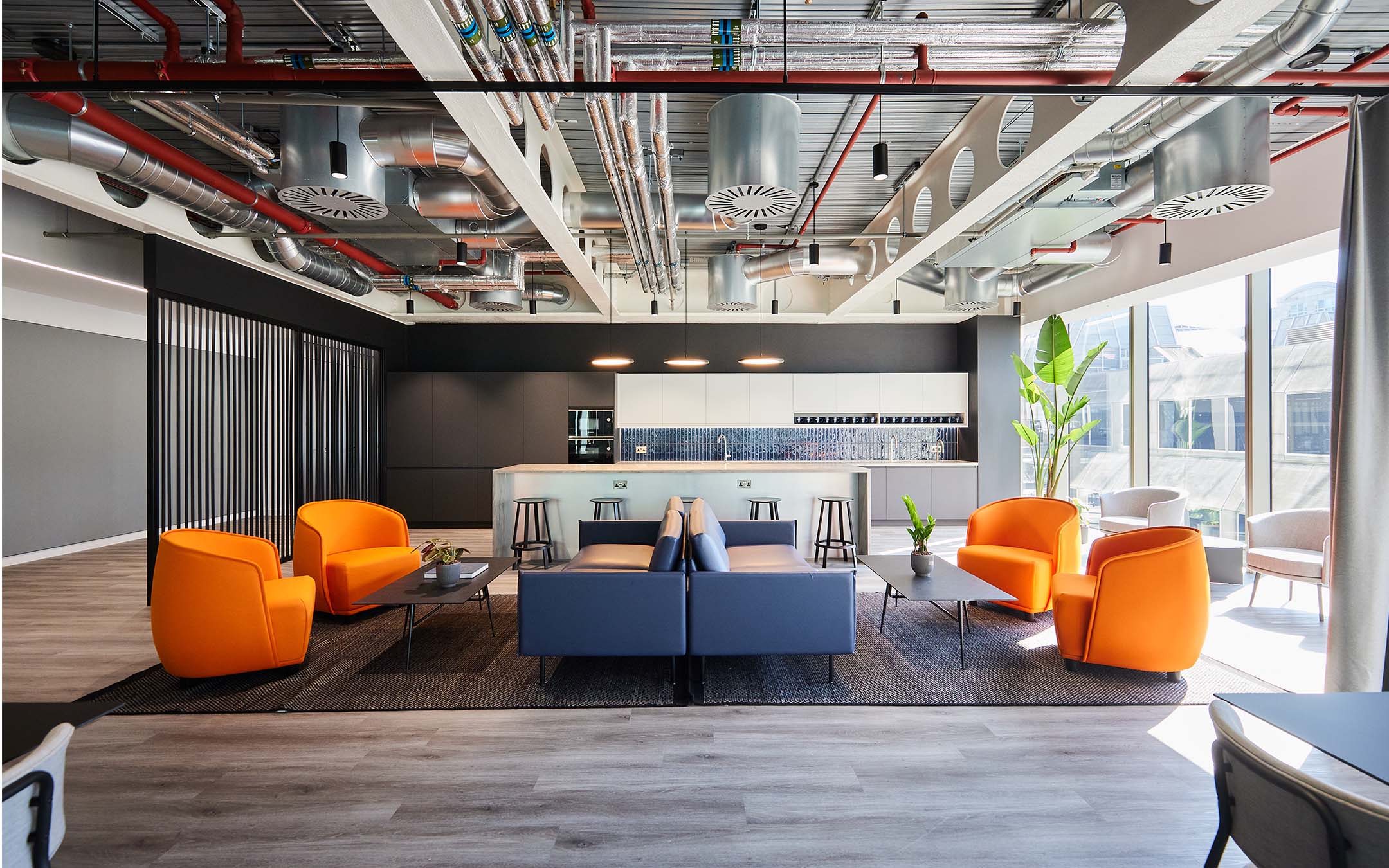 Modern office break area with orange chairs, kitchenette, and exposed ceiling.