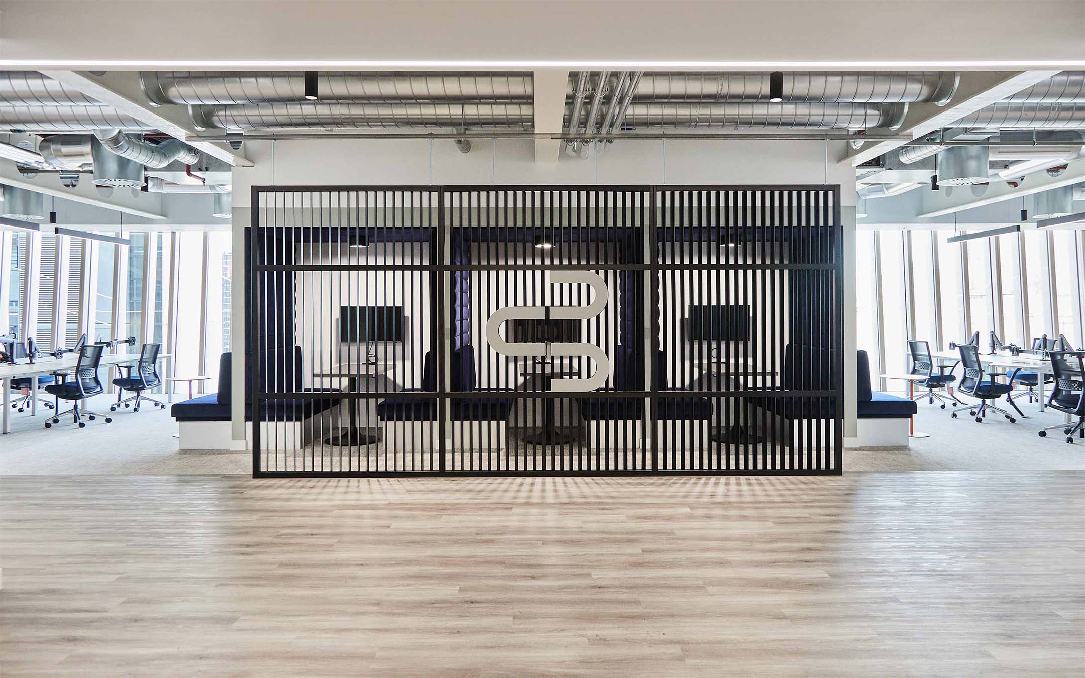 Modern office interior with desks and chairs, decorative partition with logo, and exposed ceiling.