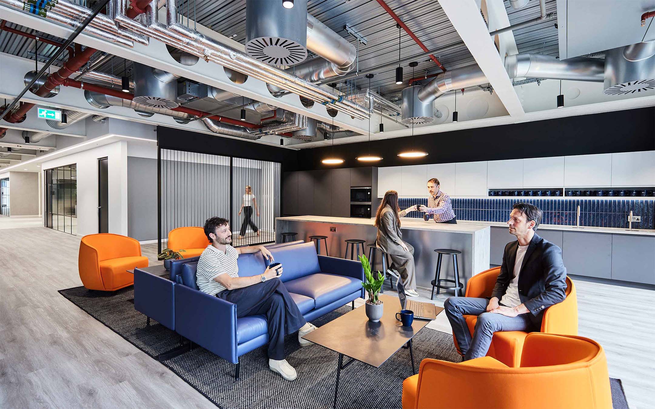 Modern office lounge area with colourful furniture and exposed ceiling pipes.