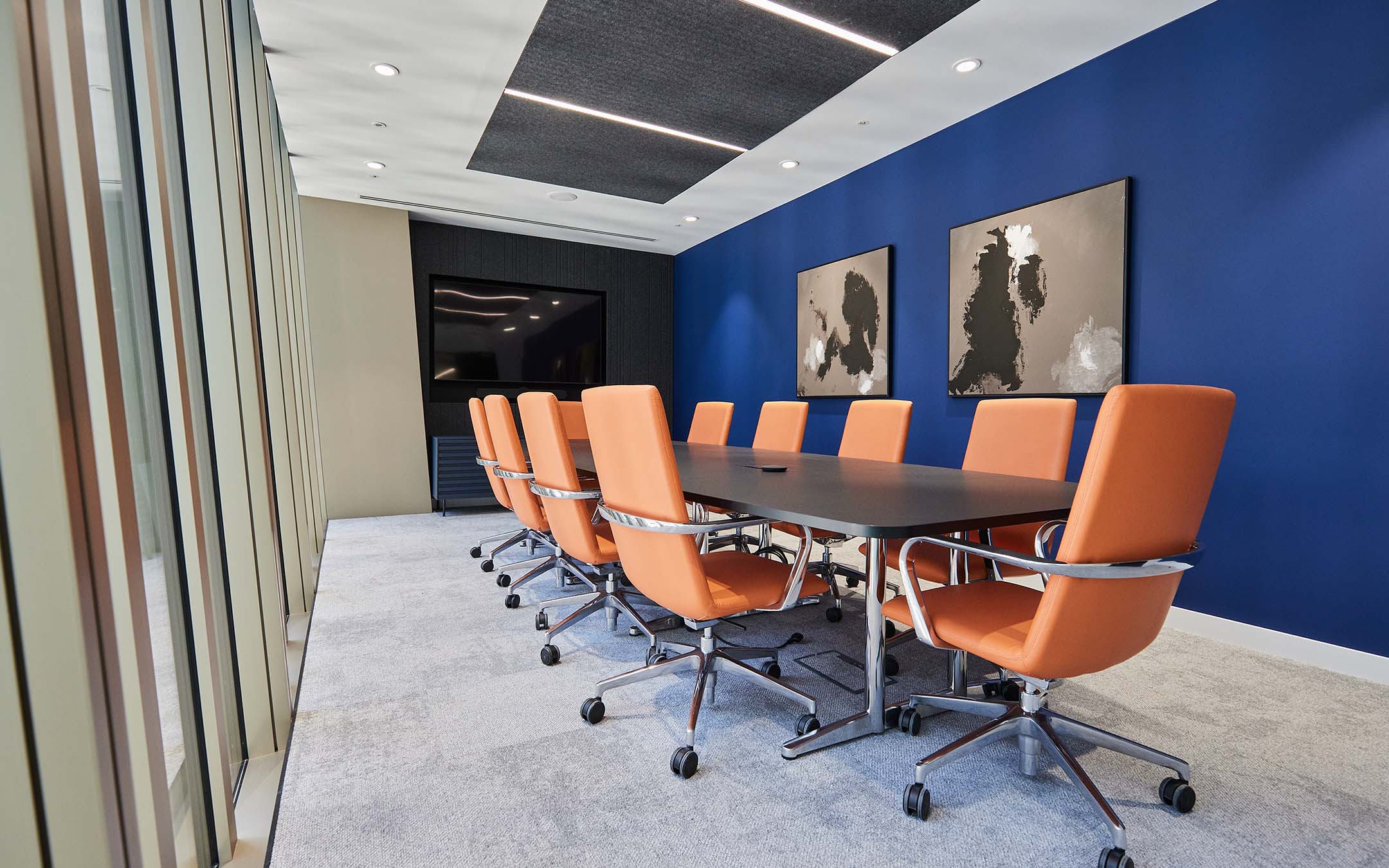 Modern boardroom with a long table, orange chairs, and blue walls adorned with art.