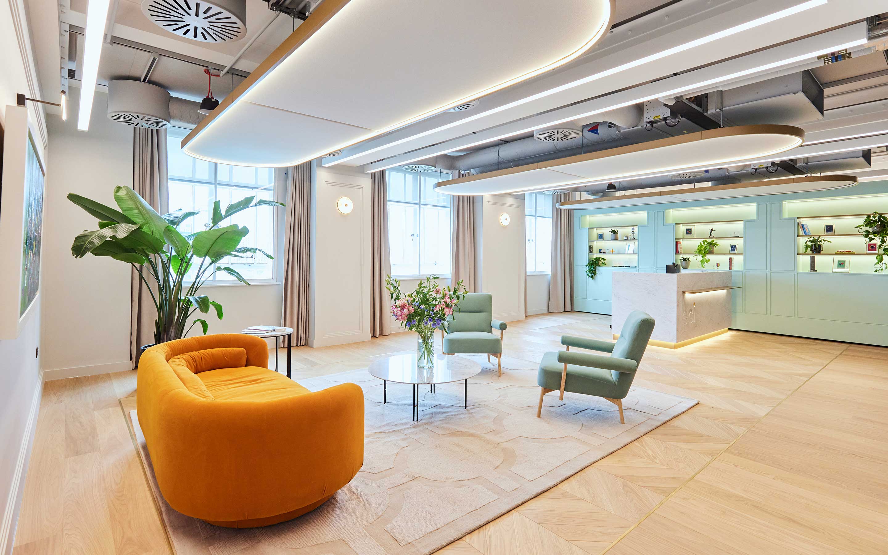 A brightly lit office reception area with a velvet yellow sofa, exposed ceiling services, and a marble reception desk