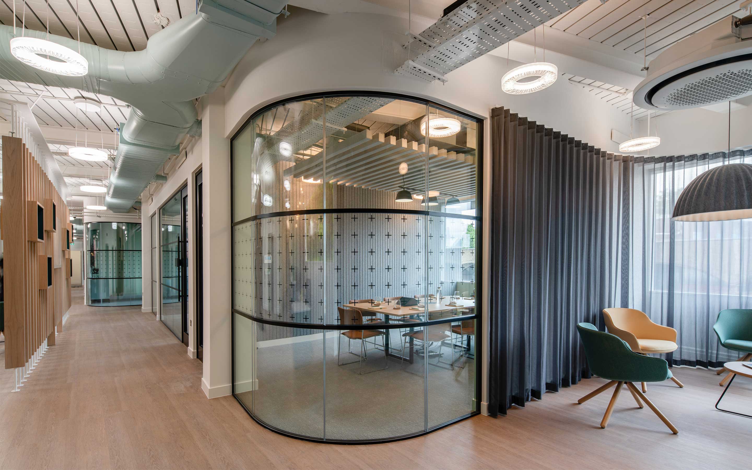 A glass walled meeting room in the middle of a modern office interior design. A soft seating area with a semi-transparent privacy curtain abuts the meeting room, creating multifunctional spaces