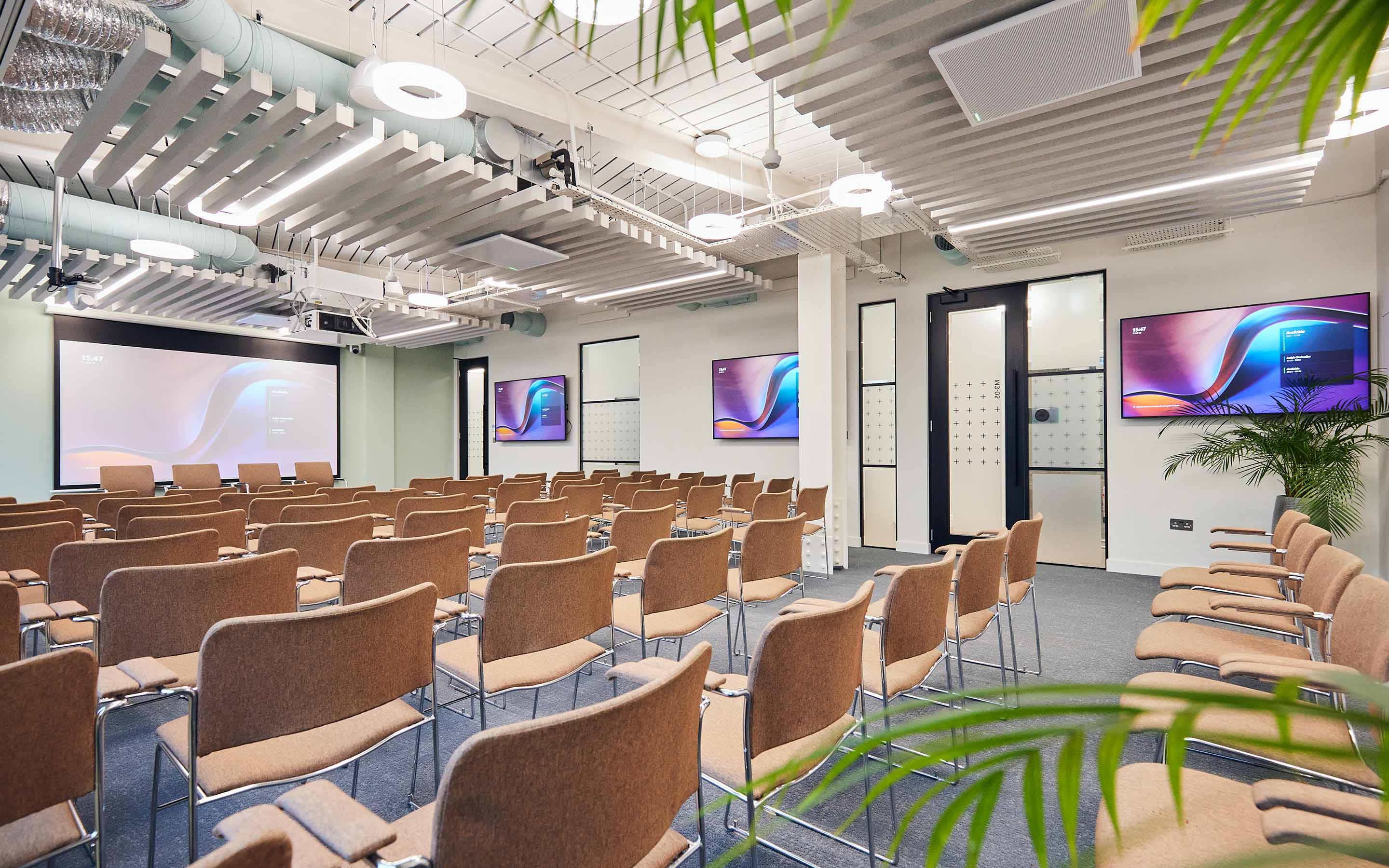 A seminar room in a contemporary office design, with pink comfortable seats, plants, and multiple AV screens