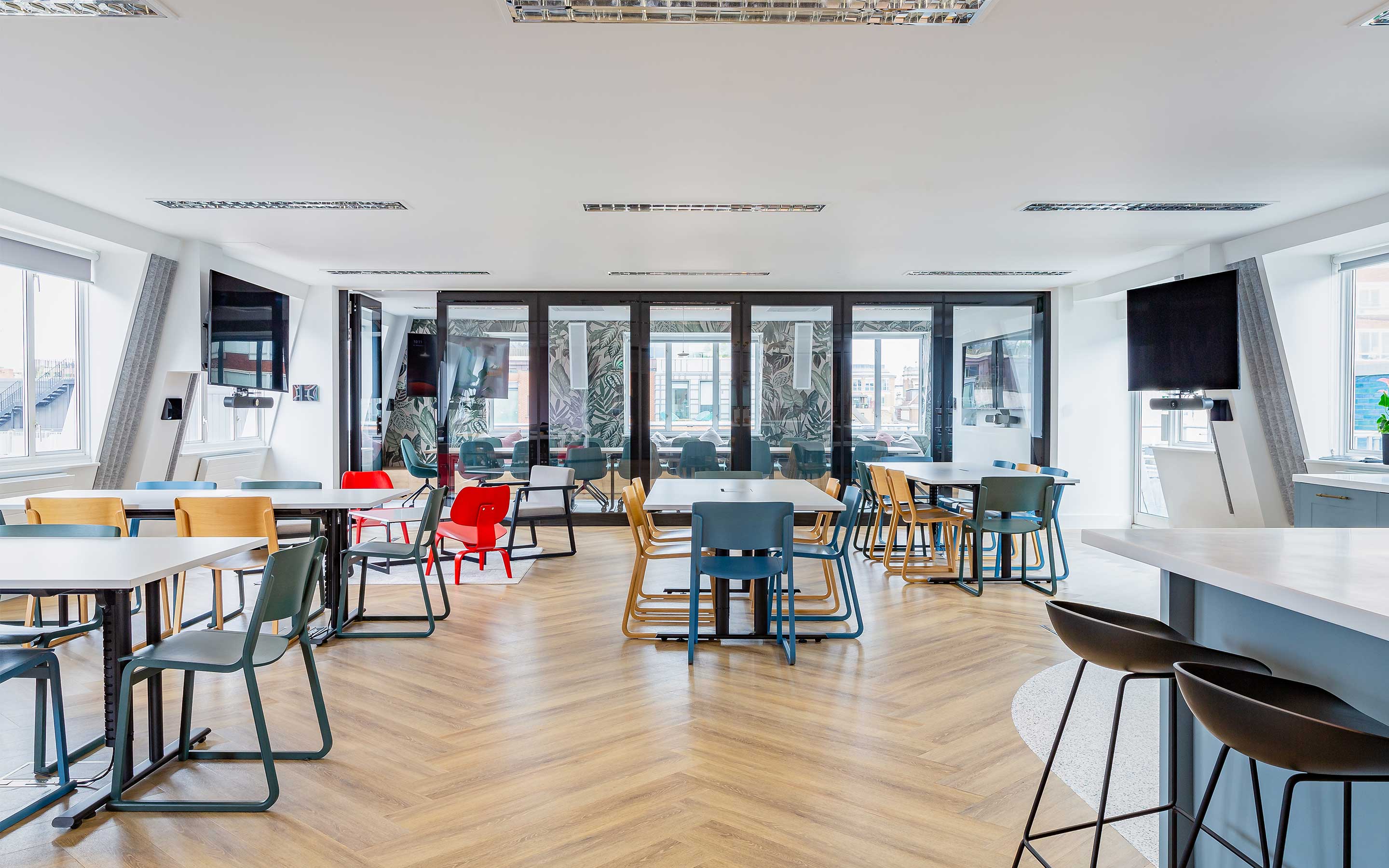 A large breakout space in a modern office interior design, with colourful chairs, wooden herringbone flooring, and lots of natural light