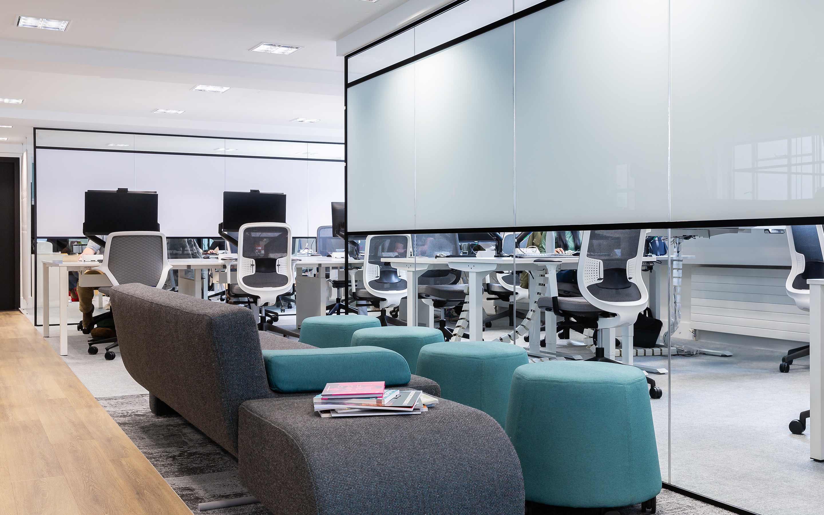 A breakout area in a modern office interior design, with glass walls, blue and grey soft seating, and views of the open place office with a clean aesthetic