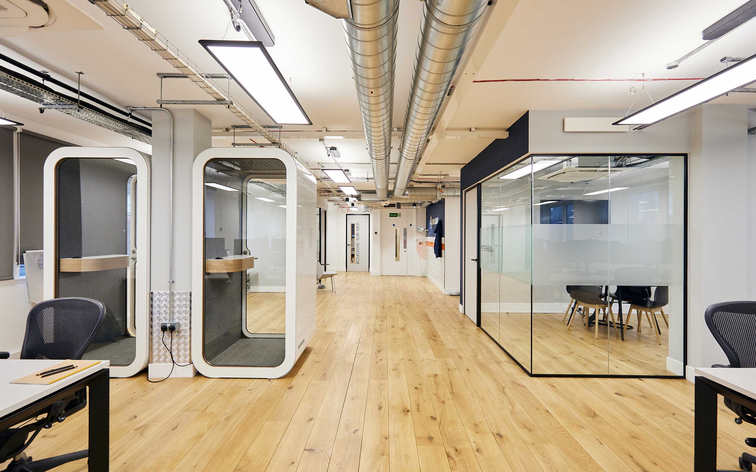A view of a sleek office interior design with glass walled meeting rooms and modern zoom booths, including wooden flooring and white walls