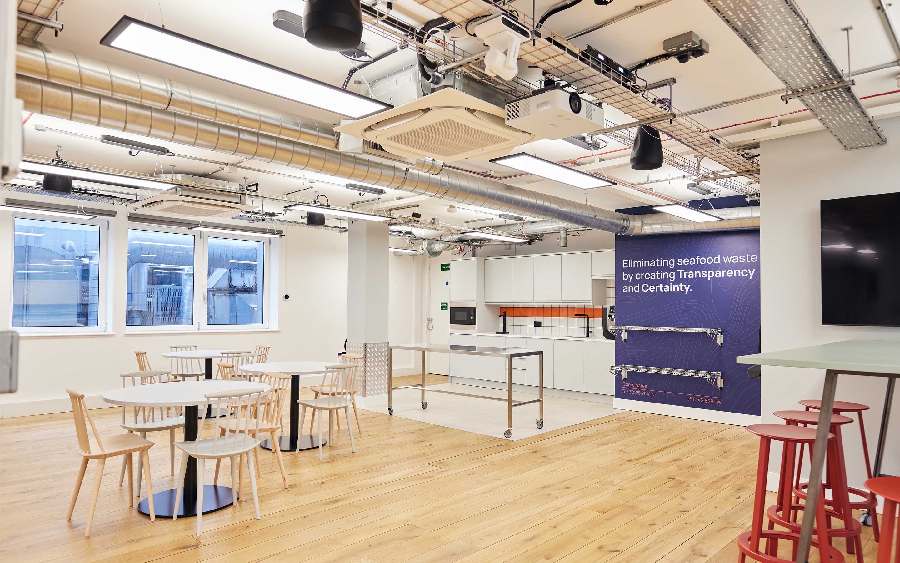 A bright office teapoint area with breakout tables, exposed ceiling services, and a navy blue manifestation wall with company slogan