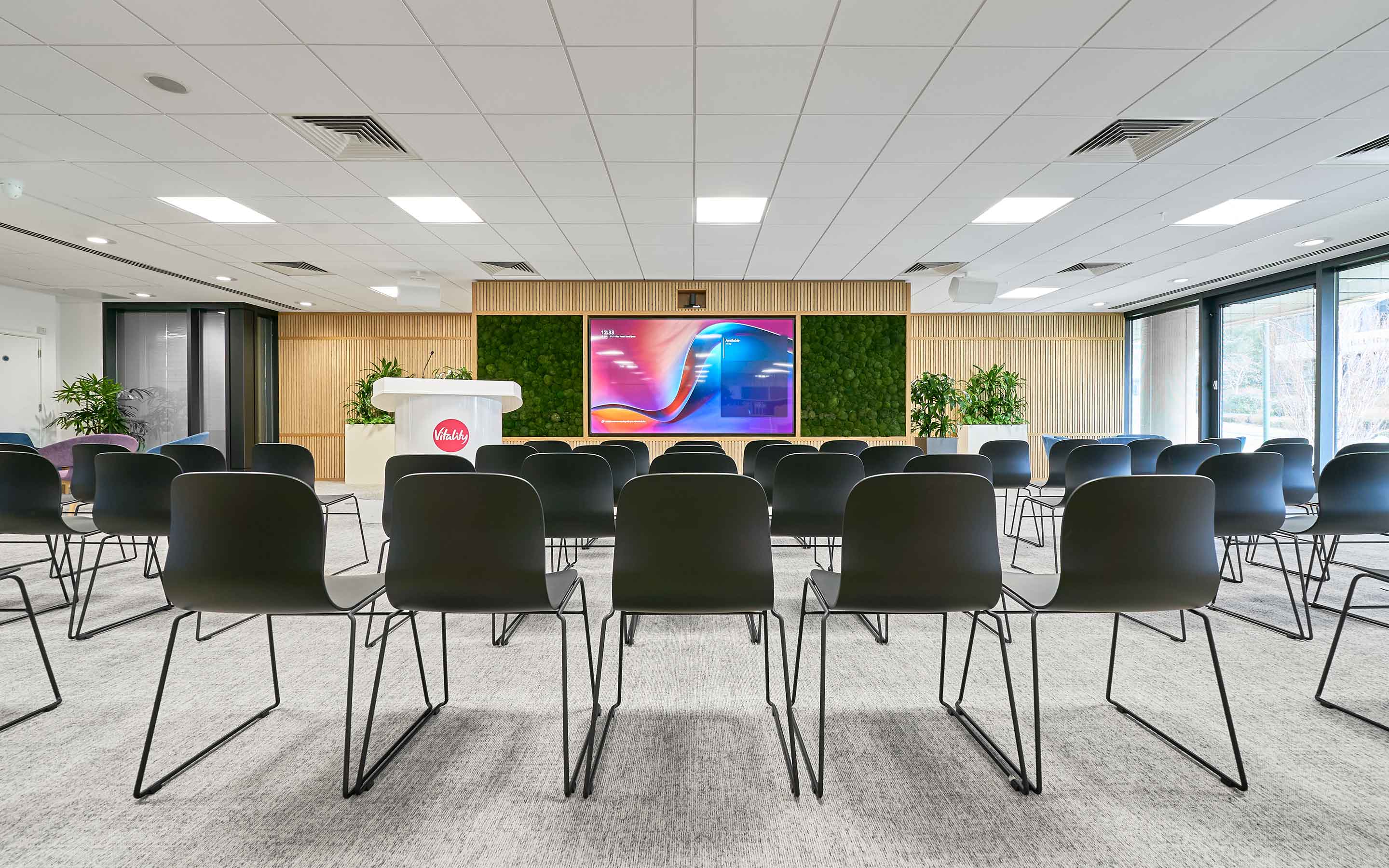 A seminar room in a modern office with chairs, a large AV screen, and podium branded to health insurer, Vitality