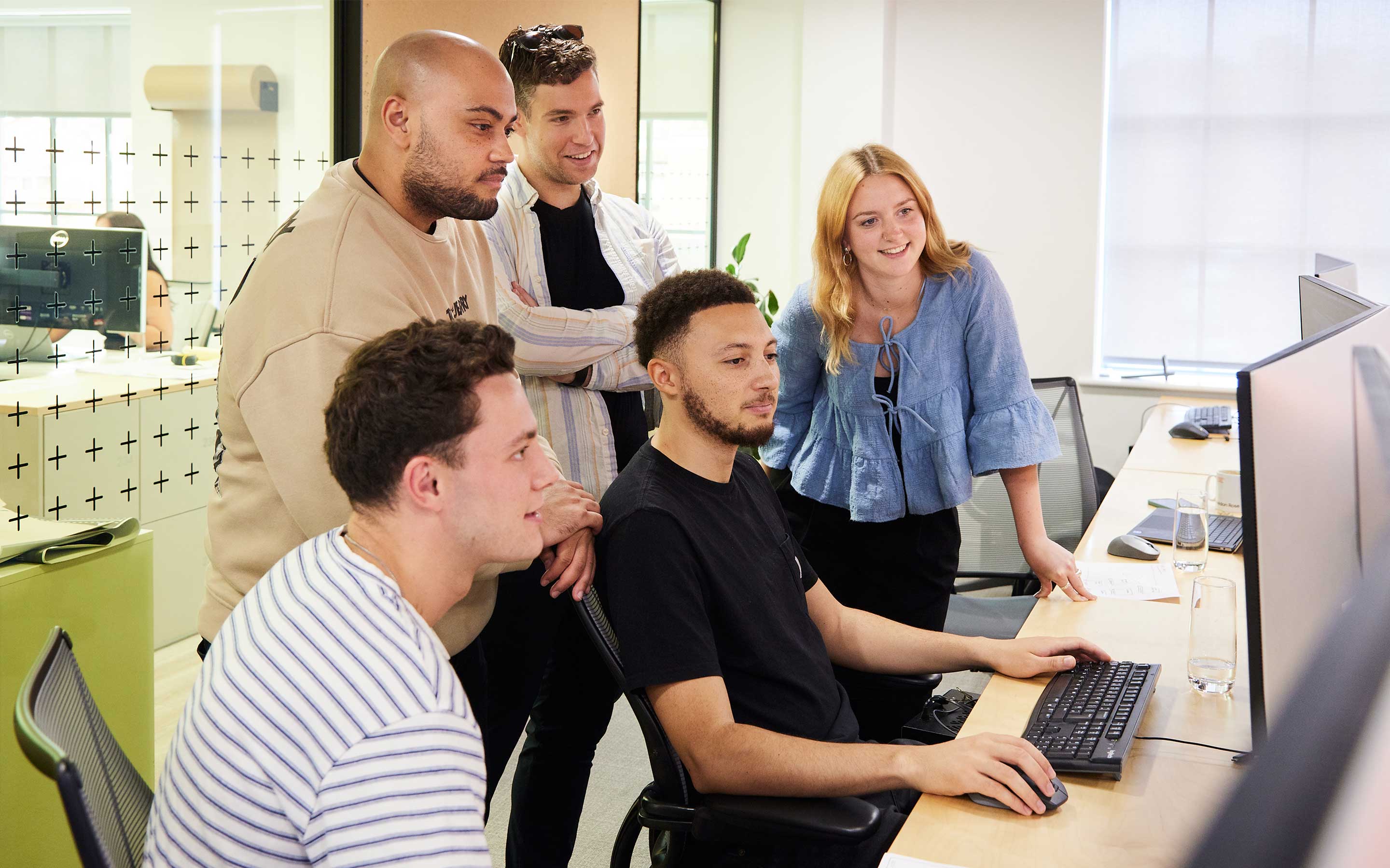 Colleagues collaborate around a computer screen smiling in a modern, agile office interior space