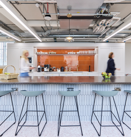 Modern office kitchenette with bar stools, two blurred figures walking by.