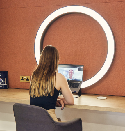 A girl on a video call facing a ring light and orange wall.