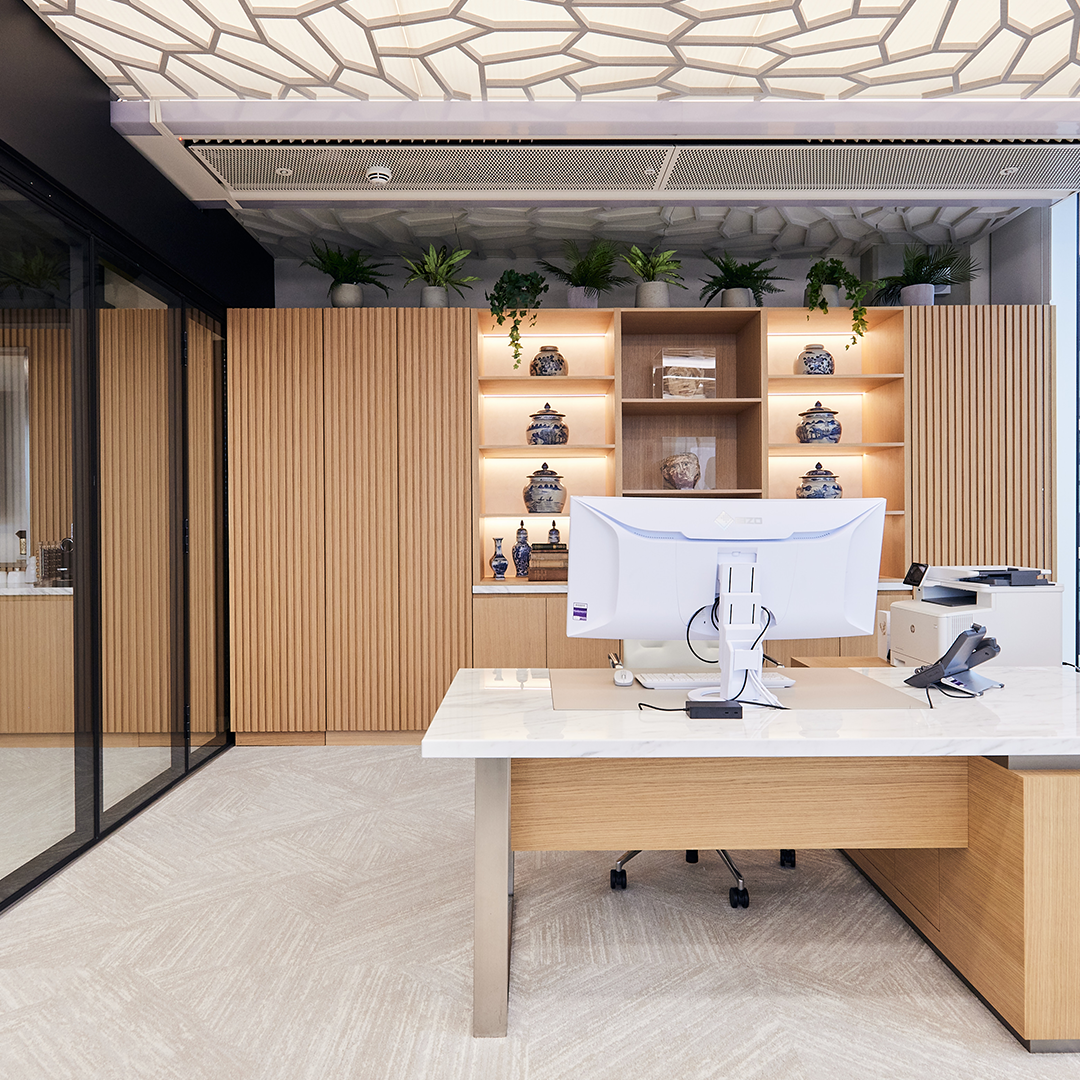 Modern office space featuring a desk with a computer, glass wall, and wooden storage cabinets adorned with plants and decorative items.