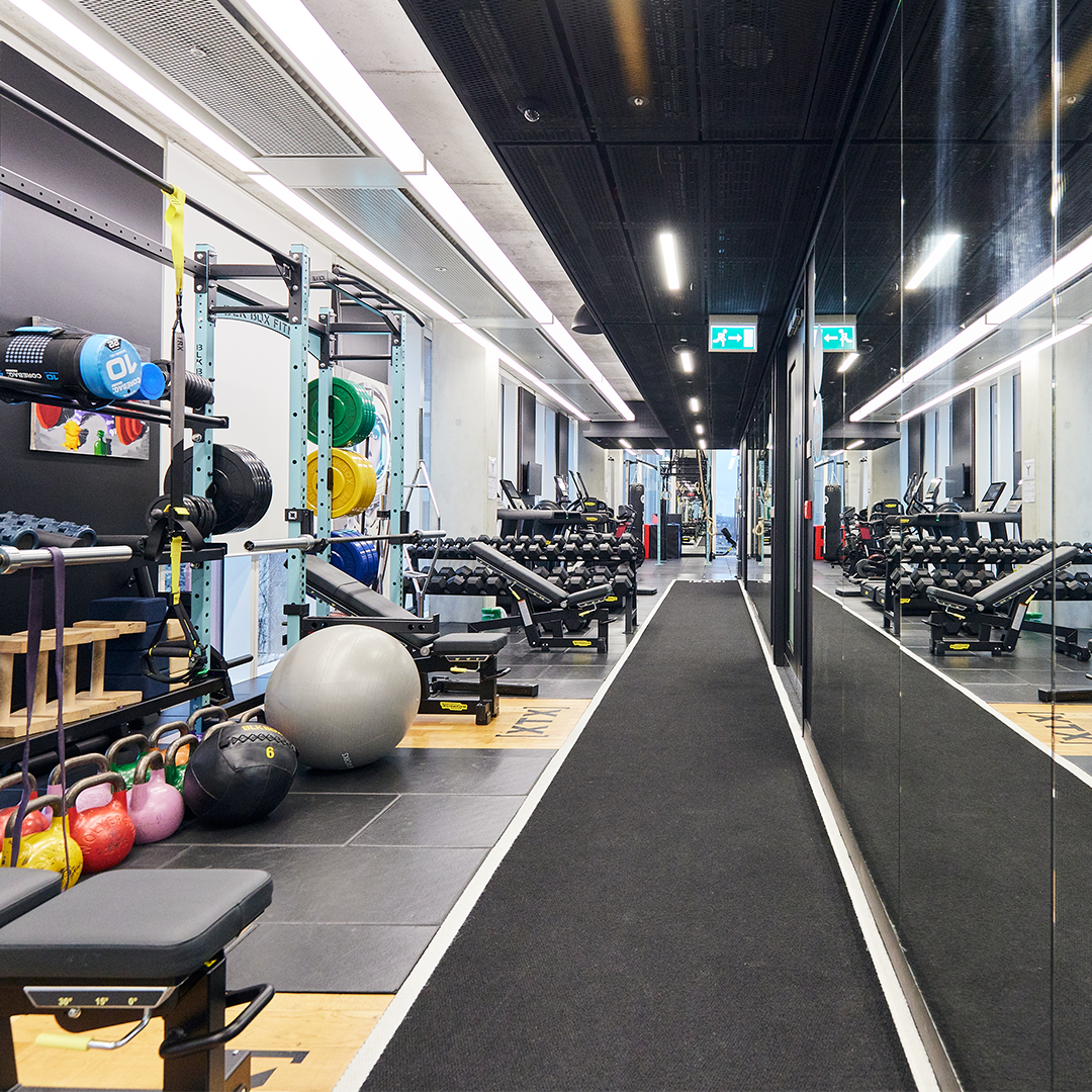 A modern gym with various equipment including weights, exercise balls, benches, and a mirrored wall reflecting the room. The space is well-lit with overhead lights and has a black floor walkway.