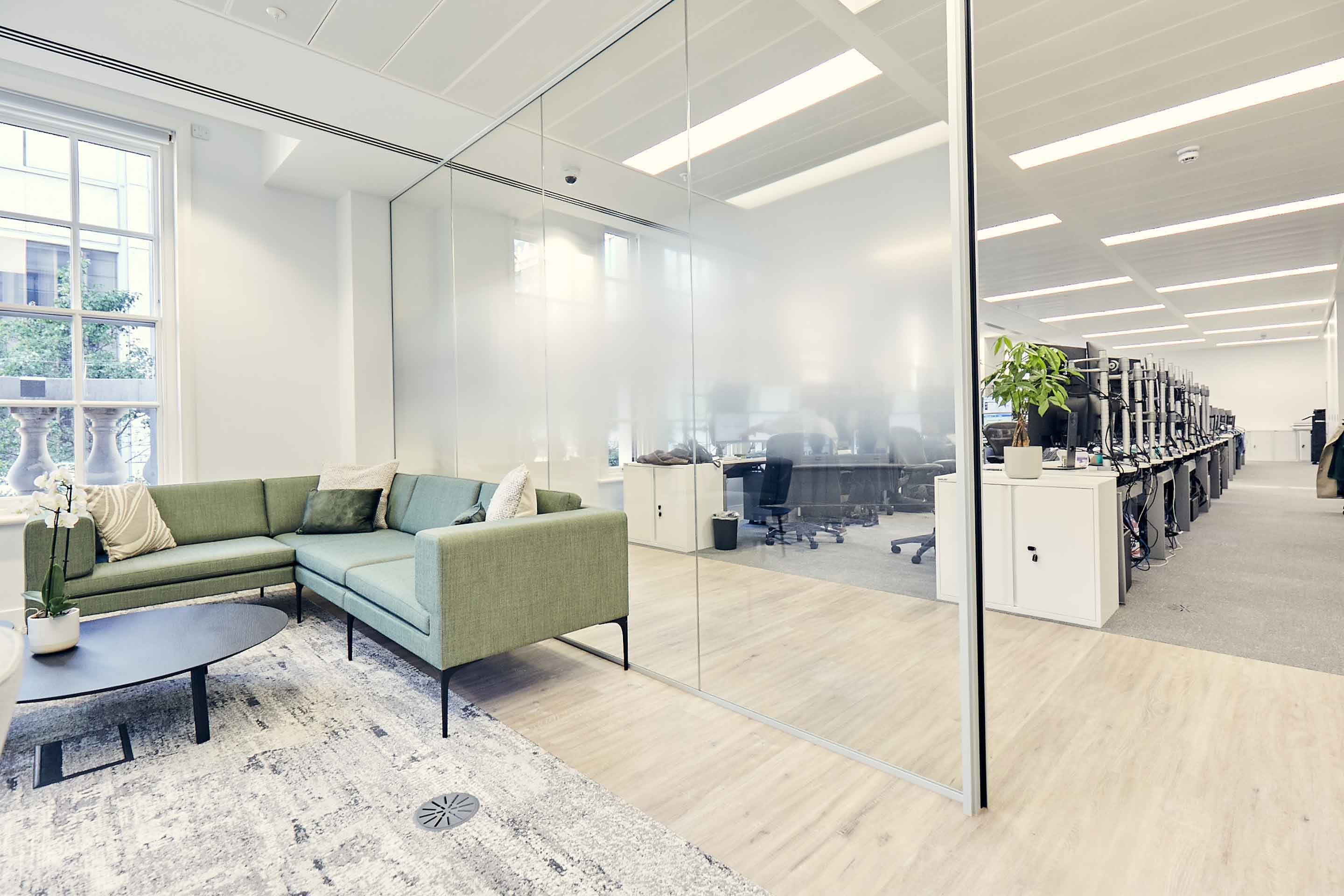 Modern office lounge with sofas beside glass partition overlooking a workspace.